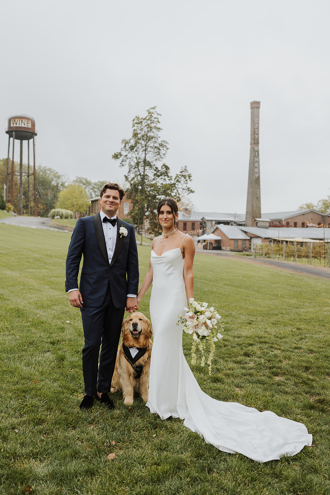 Bride and groom with their dog on the lawn at City Winery Hudson Valley, the WINE water tower and brick smokestack behind them, Montgomery NY